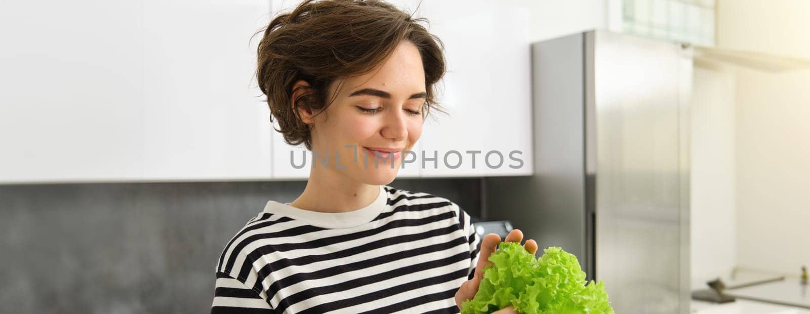 Portrait of young smiling woman in the kitchen, holding fresh green leaves of lettuce, preparing vegetables for salad, cooking healthy vegetarian meal by Benzoix