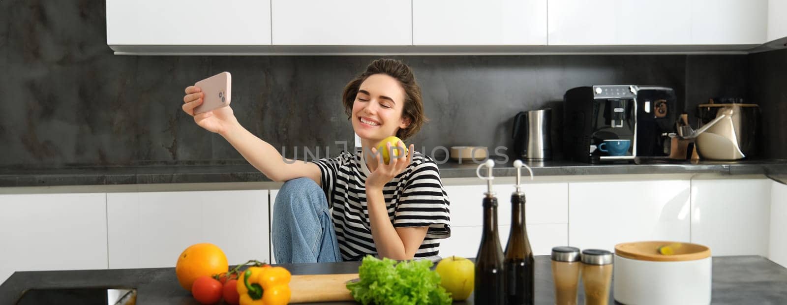 Cute young woman, modern lifestyle blogger, taking selfie while cooking salad in the kitchen, using smartphone to make photos for social media app by Benzoix