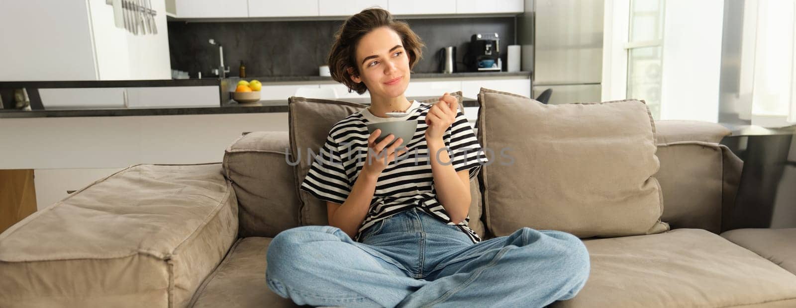 Portrait of cute young woman eating breakfast, having cereals with milk on sofa in living room, smiling and looking satisfied by Benzoix