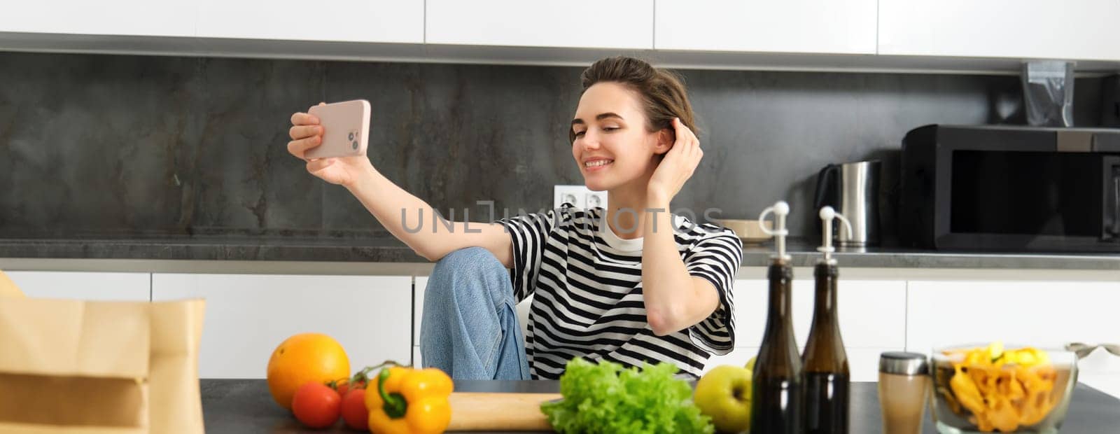 Cute young woman, modern lifestyle blogger, taking selfie while cooking salad in the kitchen, using smartphone to make photos for social media app.