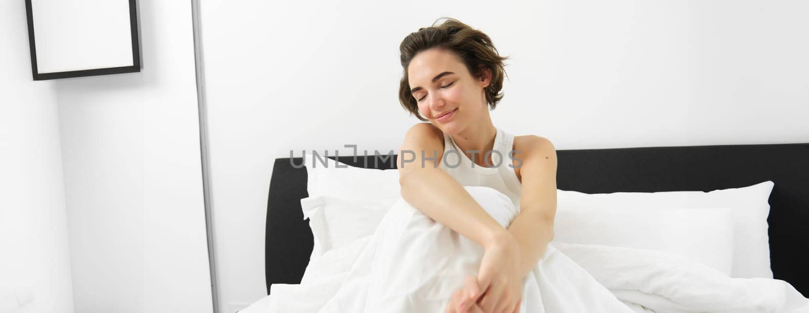 Portrait of cute smiling woman, sitting in bed, resting at home, lying under white linen sheets, waking up in morning with happy, pleased face expression by Benzoix