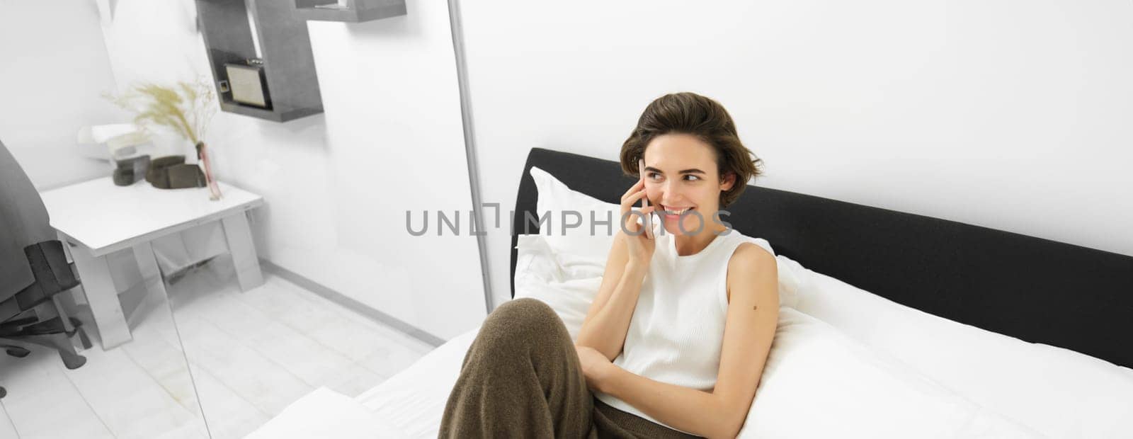 Chatty young woman laughing, talking on phone, calling friend, lying in bed and having conversation with someone, resting in bedroom by Benzoix