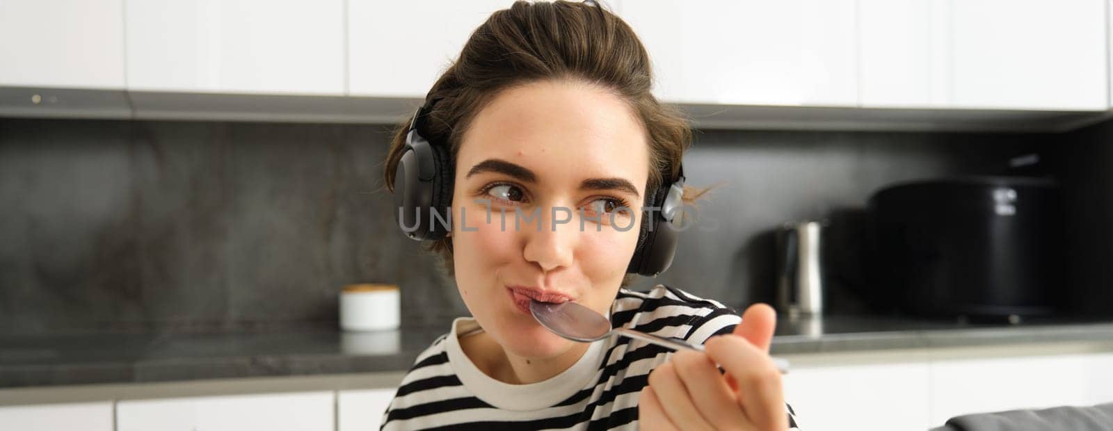 Close up portrait of young smiling woman in headphones, eating cereals with spoon and listening music, wearing earphones, sitting in the kitchen.