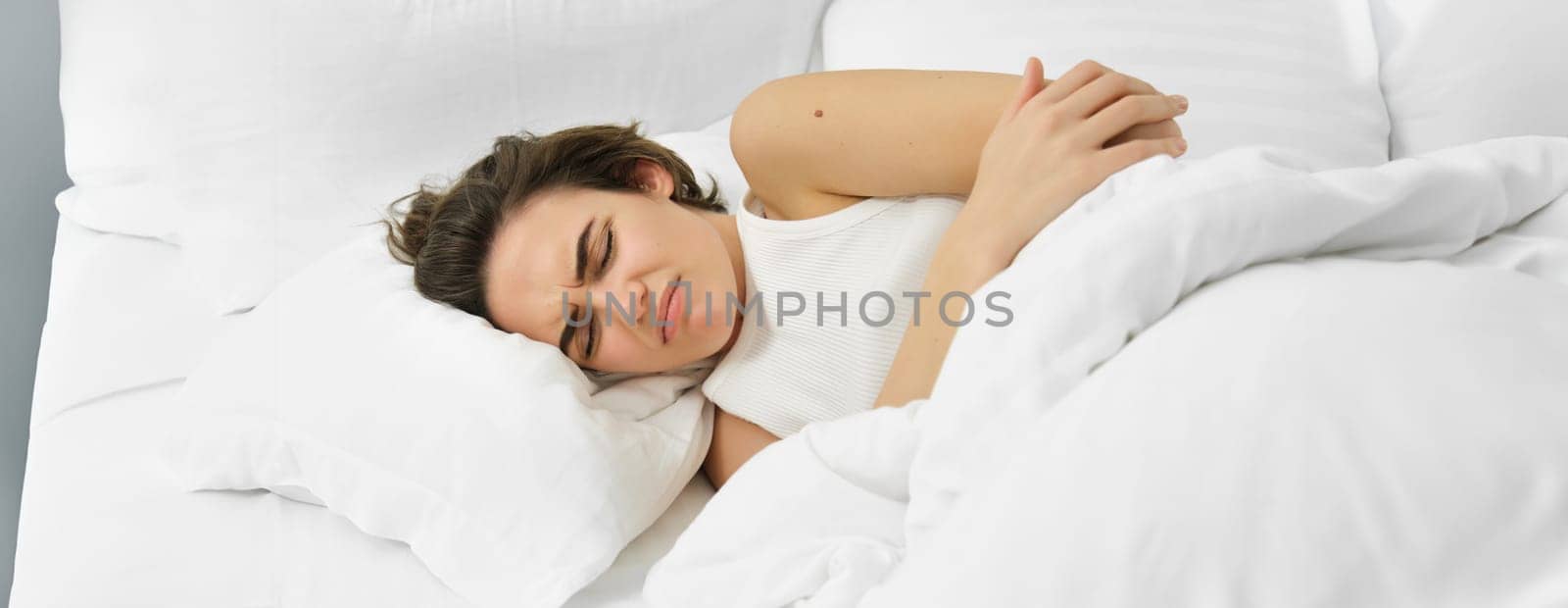 Young woman lying in bed with stomach ache, has painful cramps, menstrual pain. Lifestyle and wellbeing concept