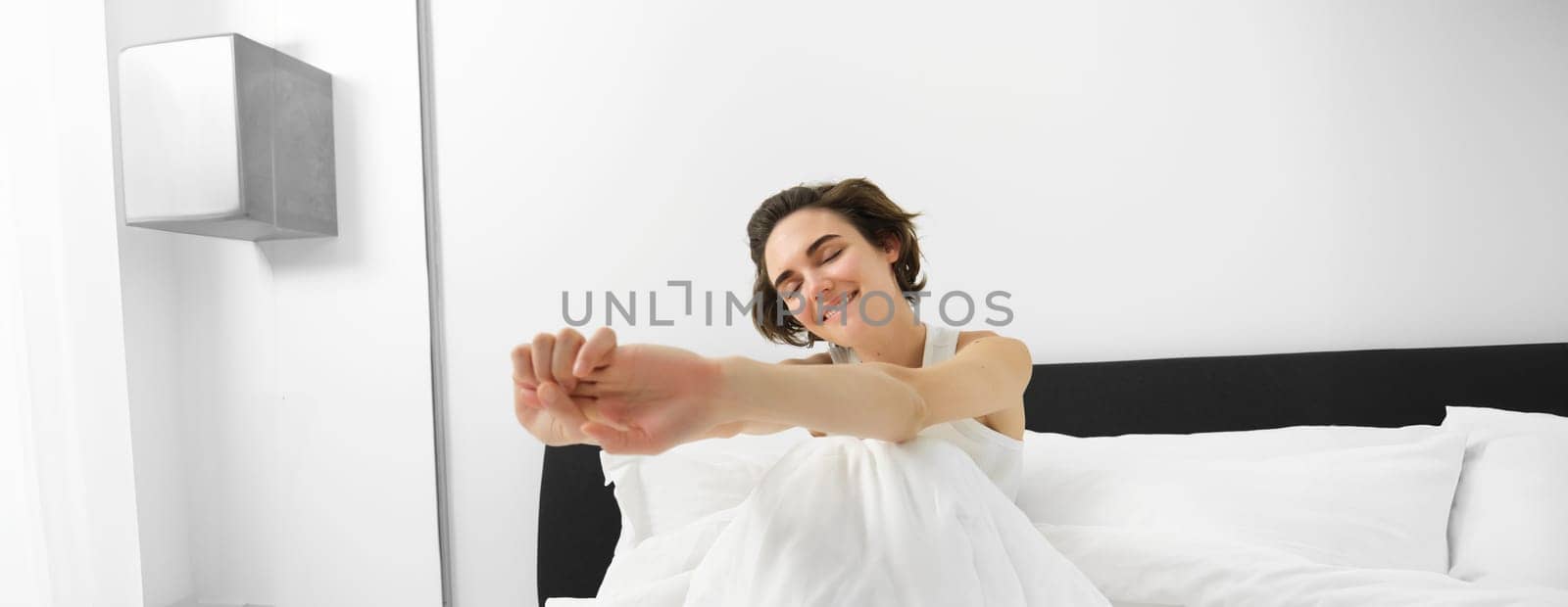 Portrait of smiling young woman wakes up in her bed, sitting under white linen sheets with closed eyes and stretching her hands with satisfaction.