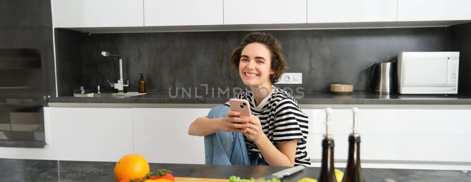 Portrait of young woman searching for cooking recipes online using smartphone, sitting near vegetables, salad ingredients and chopping board, smiling at camera.