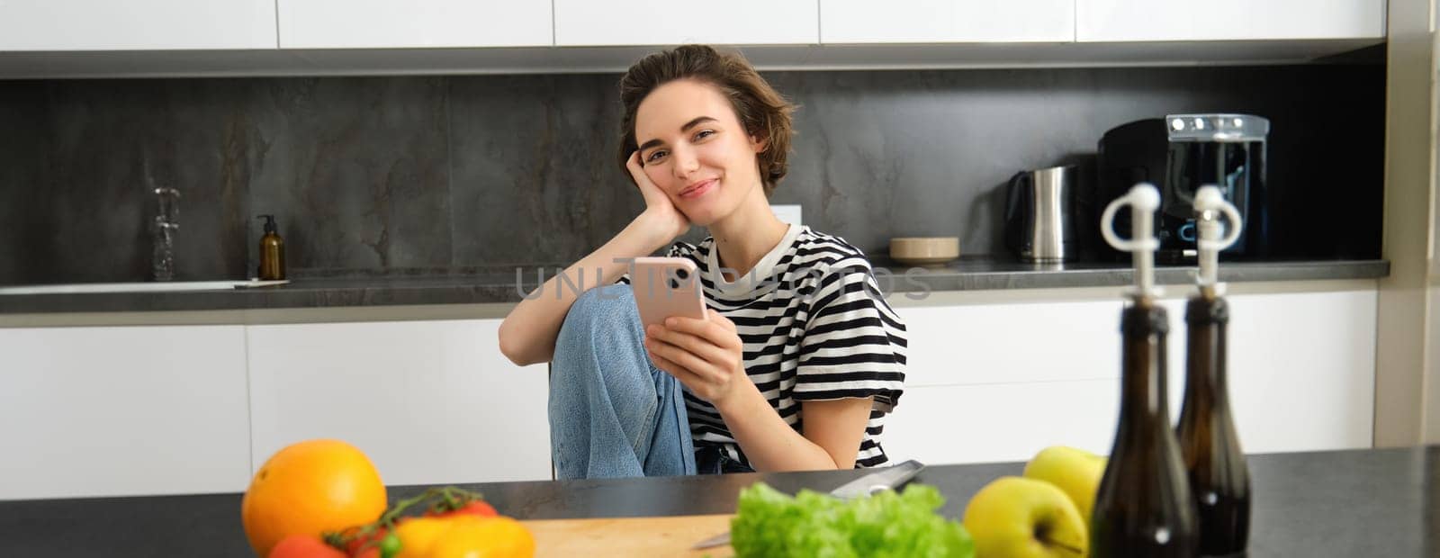 Portrait of young woman searching for cooking recipes online using smartphone, sitting near vegetables, salad ingredients and chopping board, smiling at camera.