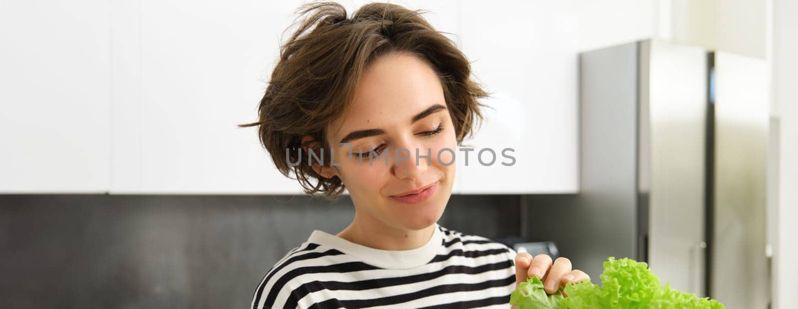 Close up portrait of smiling, beautiful female model, making a salad, holding green lettuce leaf, preparing vegetables, cooking in the kitchen, standing in striped t-shirt.