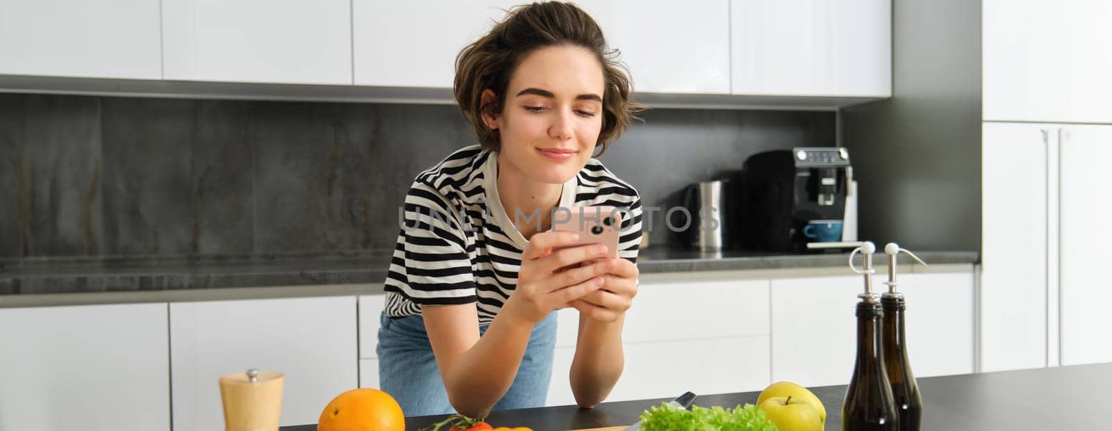 Portrait of young smiling woman, searching cooking recipe online on smartphone, standing near vegetables and chopping board, making meal, preparing salad and using social media on mobile phone by Benzoix