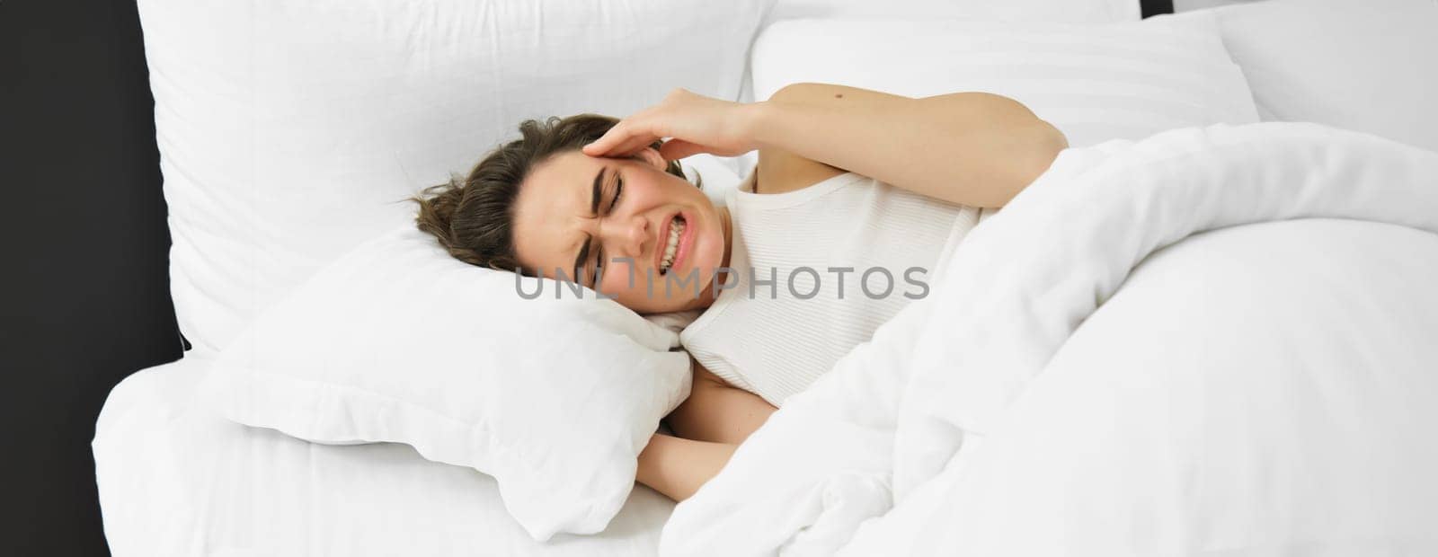 Close up portrait of female model, lying in bed with headache, grimacing and frowning from painful migraine, has pain in head, feeling unwell. Health and women concept
