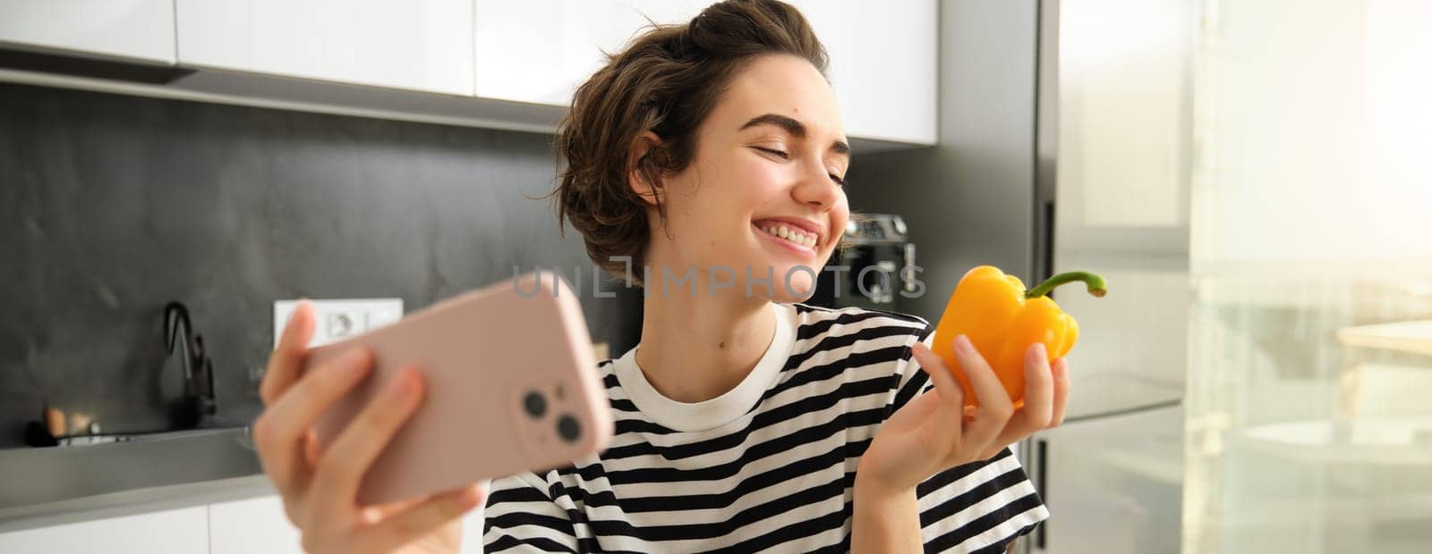 Smiling modern woman, vegetarian food blogger, taking selfie with fresh yellow sweet pepper, holding smartphone, recording a video blog for her social media account, posing in the kitchen.