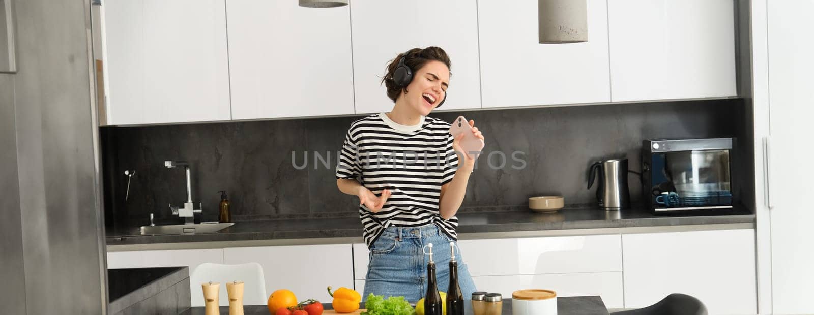 Carefree woman cooking in the kitchen, singing along favourite song, listening music in wireless headphones, holding mobile phone, chopping vegetables, preparing healthy meal.