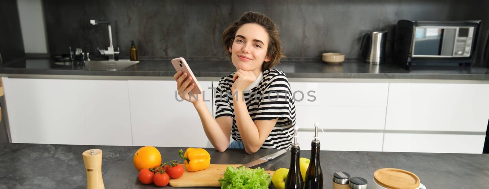 Portrait of modern young woman thinking what to cook, sitting in the kitchen with smartphone and vegetables, making a meal, healthy diet salad.