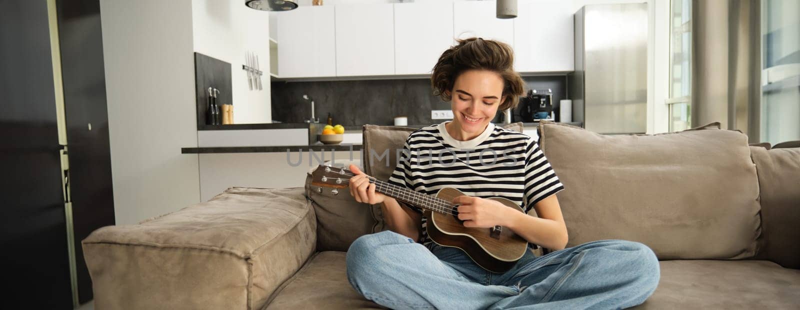 Portrait of cheerful young woman playing her ukulele, singing and laughing, sitting in living room at home by Benzoix