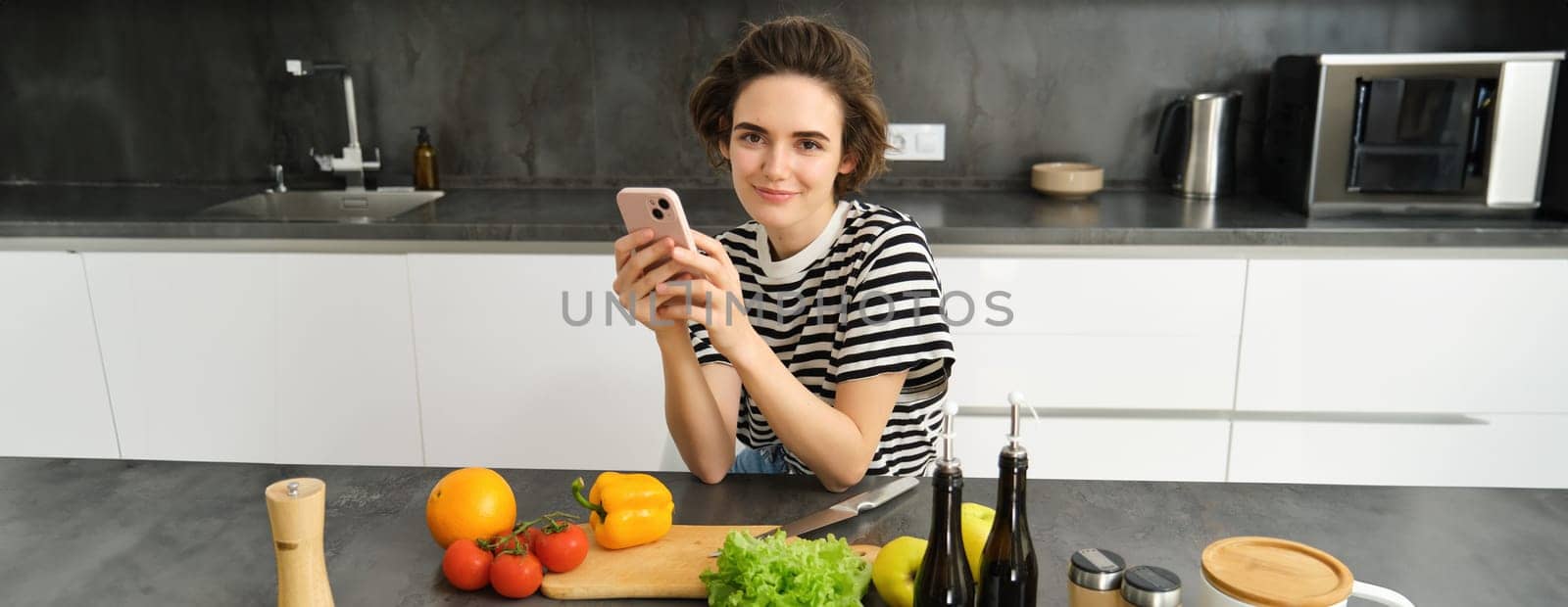 Portrait of modern young woman thinking what to cook, sitting in the kitchen with smartphone and vegetables, making a meal, healthy diet salad.