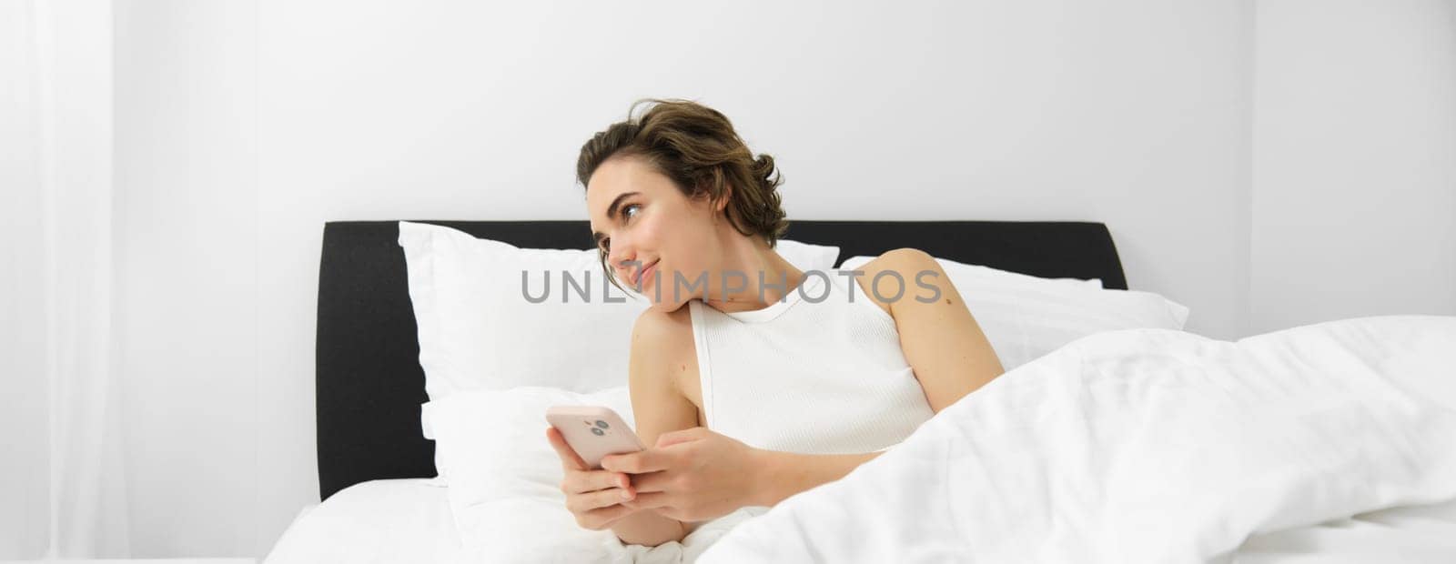 Portrait of woman in bed, lying on white sheets, using smartphone, holding mobile phone. Lifestyle and technology concept