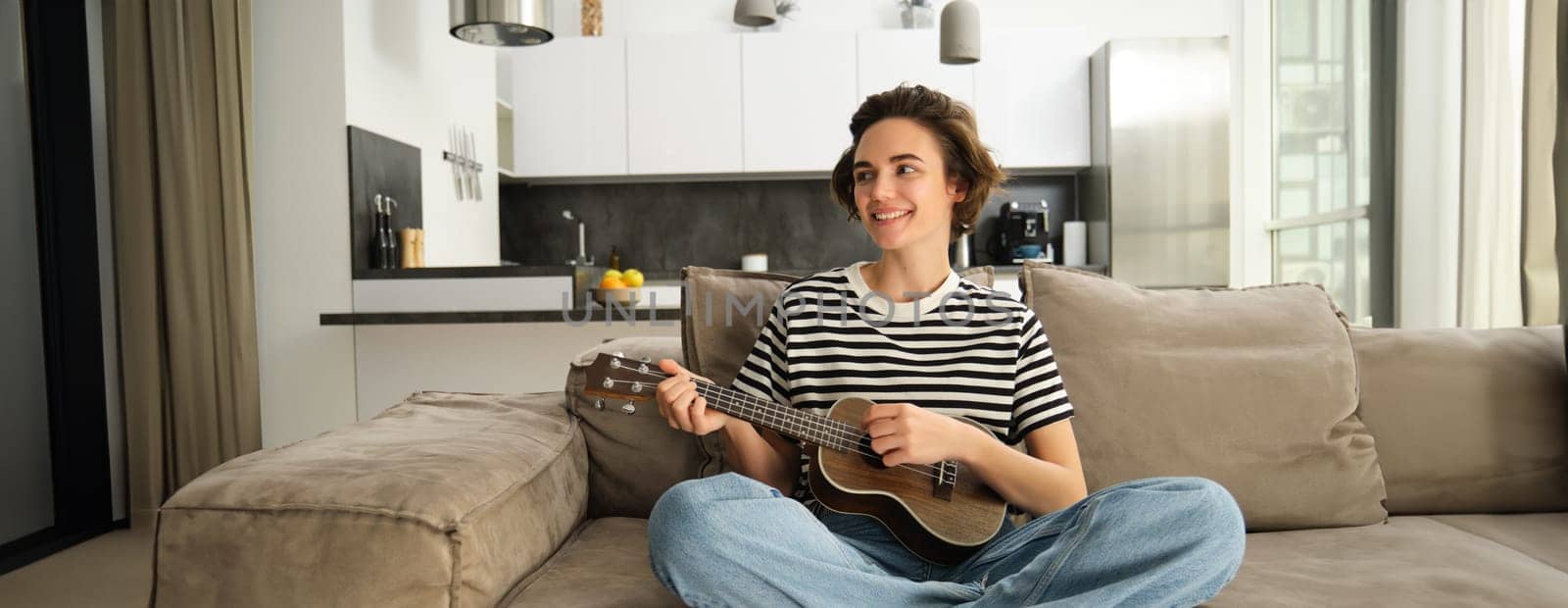 Portrait of cute young woman sitting on sofa, learns how to play ukulele, holding her music instrument, picking chords, resting in living room.