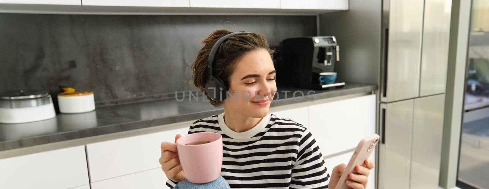 Close up portrait of young female student, drinking tea and listening to music in headphones, using smartphone in kitchen.