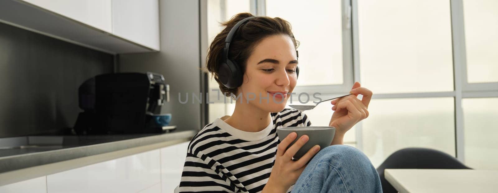 Close up portrait of smiling, beautiful young woman in headphones, eating cereals for breakfast and listening music or e-book, holding spoon with bowl.