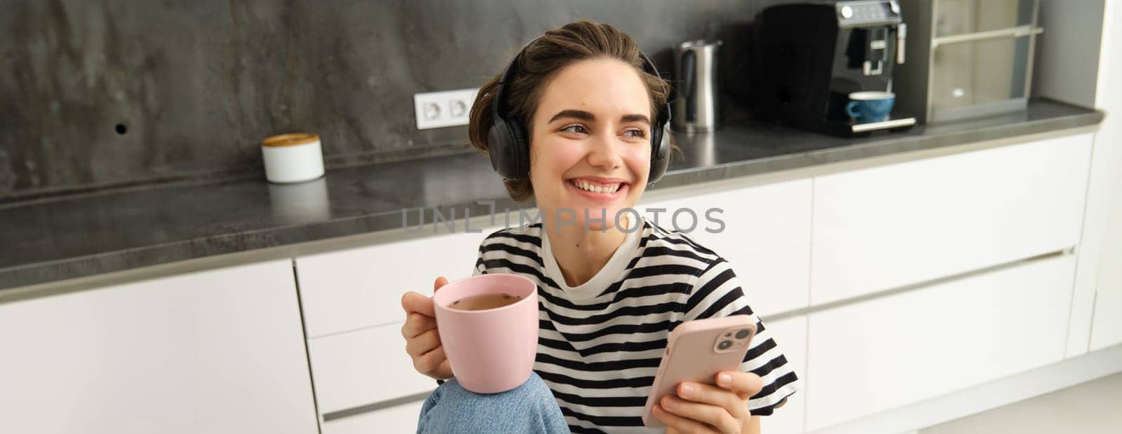 Stylish young woman in headphones, listens music and uses smartphone, drinks warm tea in kitchen, relaxing at home by Benzoix