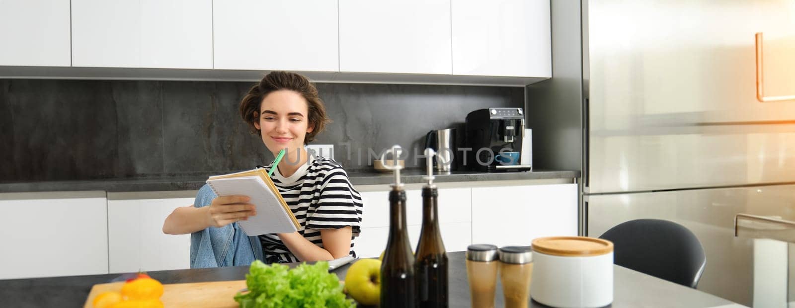 Portrait of young woman looking at cooking ingredients on kitchen counter and making notes, writing down recipes, thinking of meal for dinner, preparing vegetarian food.