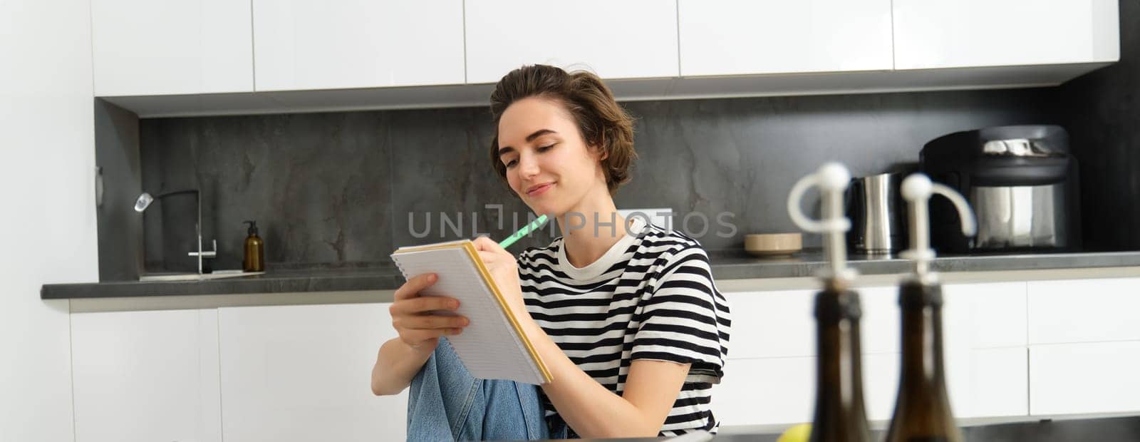 Portrait of beautiful modern woman, girl cooking with vegetables, holding notebook, writing down recipe, making notes, dinner plans, creating meal list for weekend, sitting in kitchen.