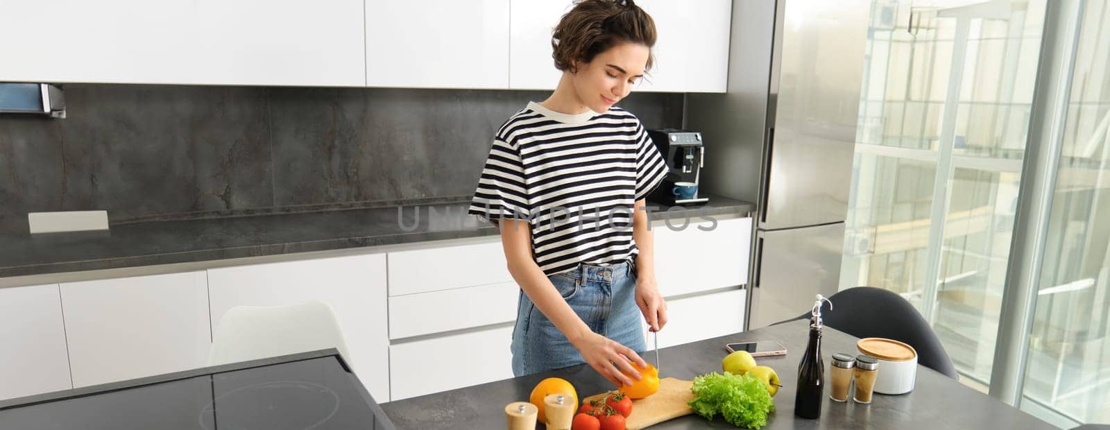 Portrait of modern young woman standing in the kitchen with vegetables, chopping them on counter, holding knife, cooking meal, vegetarian food, concept of healthy lifestyle an diet.