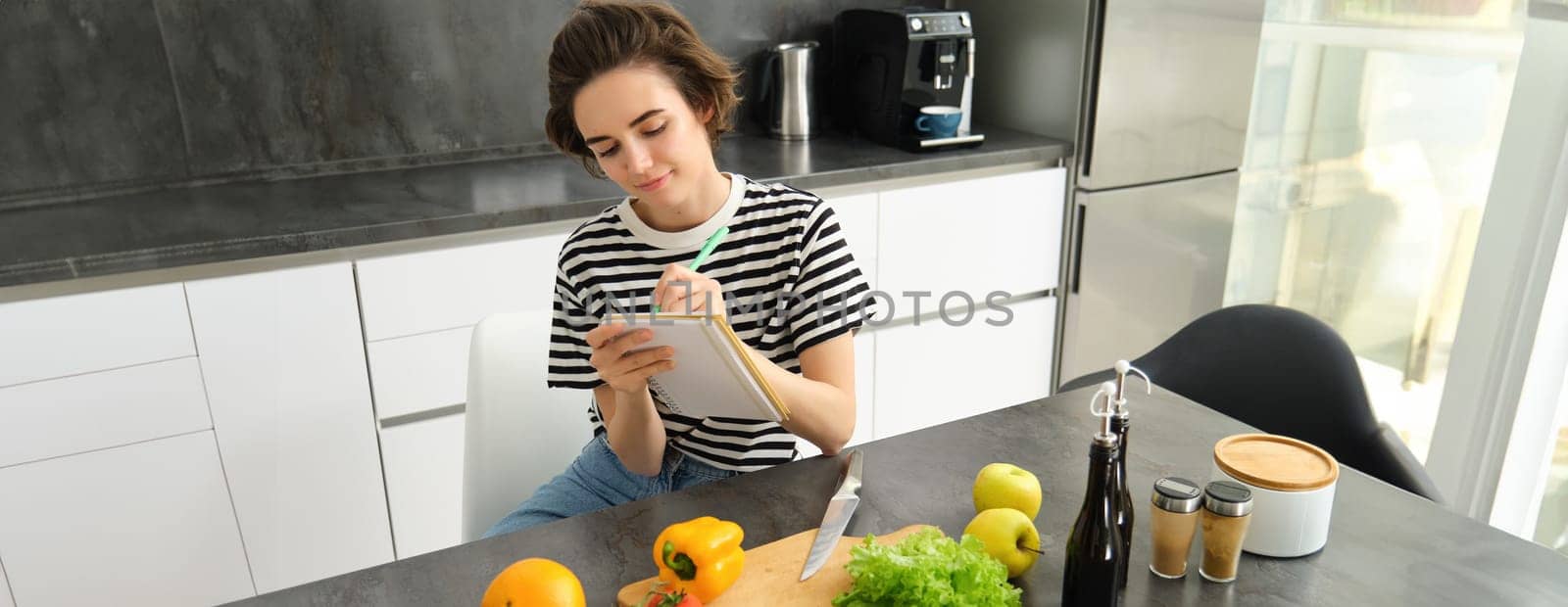 Young brunette woman writing down recipe, making notes in notebook while cooking a salad, thinking of grocery list, sitting in the kitchen with vegetables on the table by Benzoix