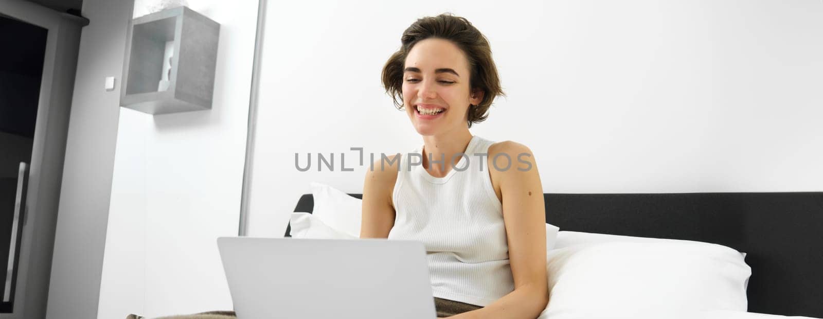 Modern woman resting at home, sitting on bed with laptop, looking at computer screen, laughing and smiling.