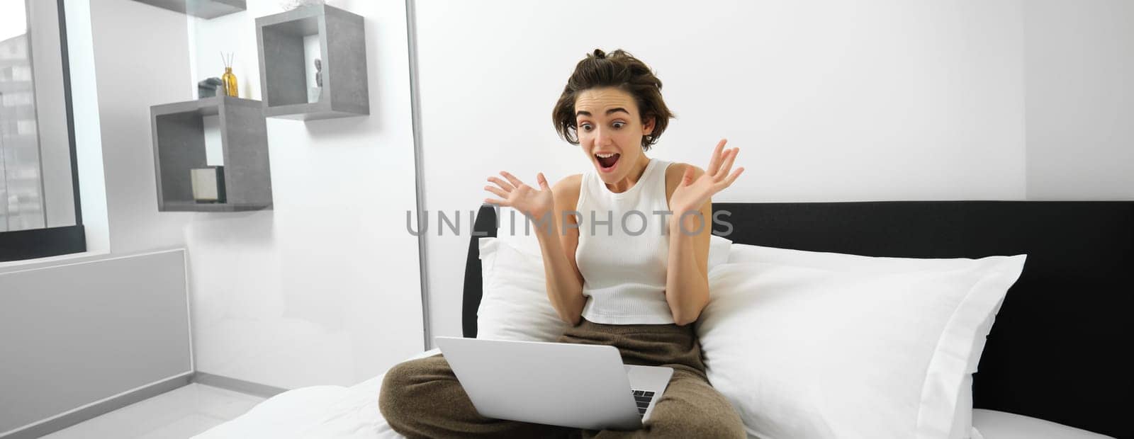 Image of woman sitting on bed, looking at smartphone with surprised, shocked face expression, has amazed reaction to online deal by Benzoix