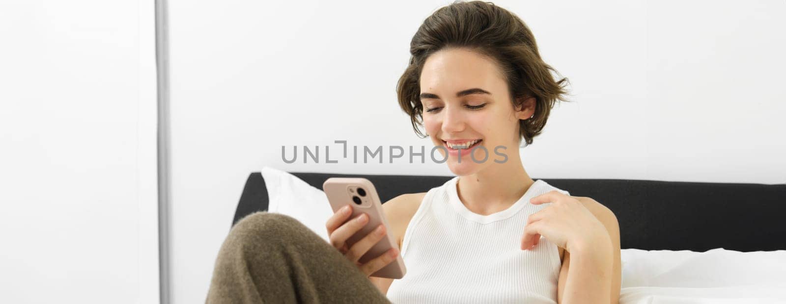 Beautiful woman lying in bed with smartphone, resting in her hotel suite, using mobile phone for food delivery, reading on cellphone by Benzoix