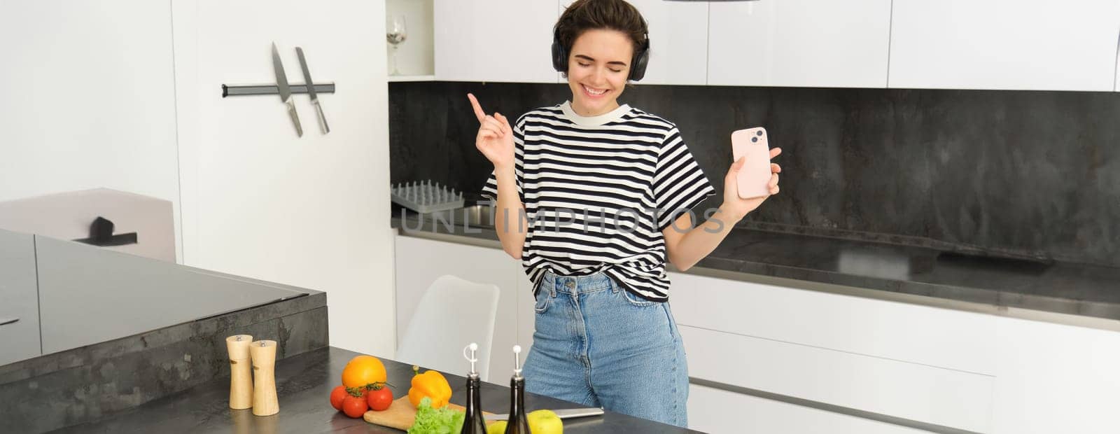 Portrait of happy dancing woman, cooking food in the kitchen, listening music in wireless headphones, chopping vegetables for salad, preparing healthy food and enjoying favourite song by Benzoix