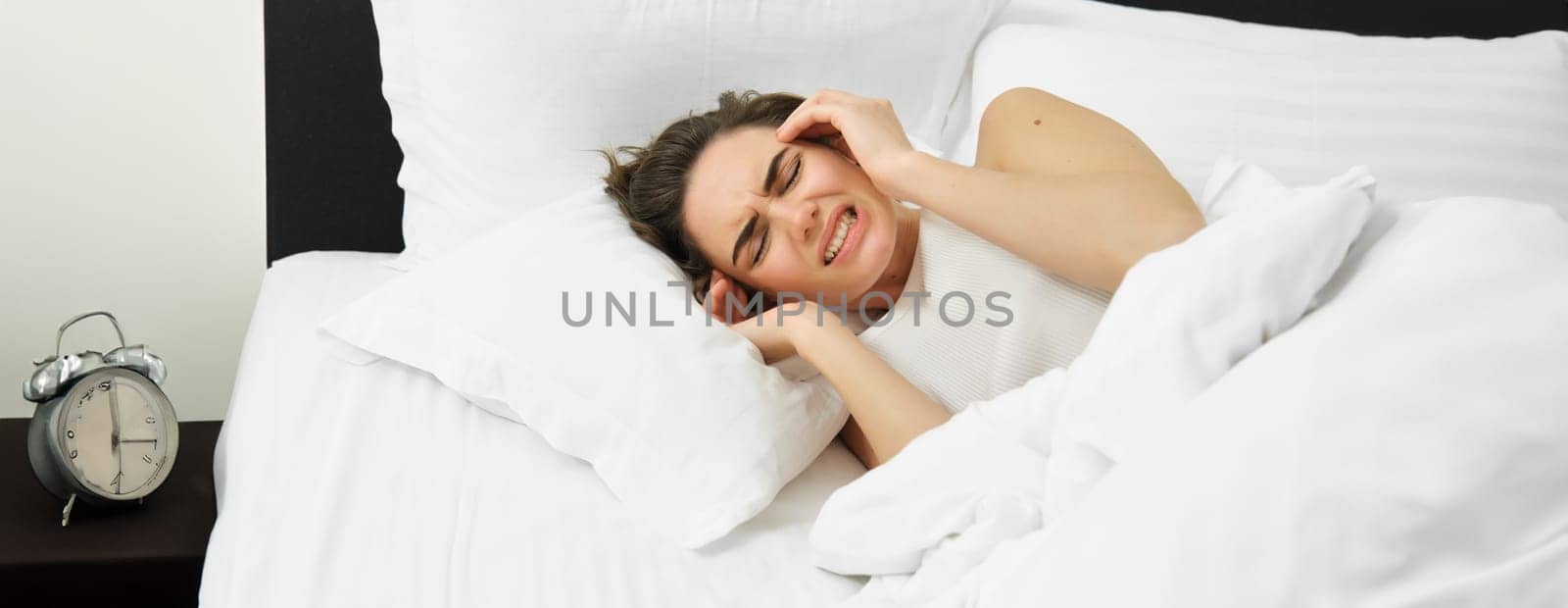 Portrait of woman with headache, lying in bed and suffering from migraine, touching head and grimacing from discomfort.