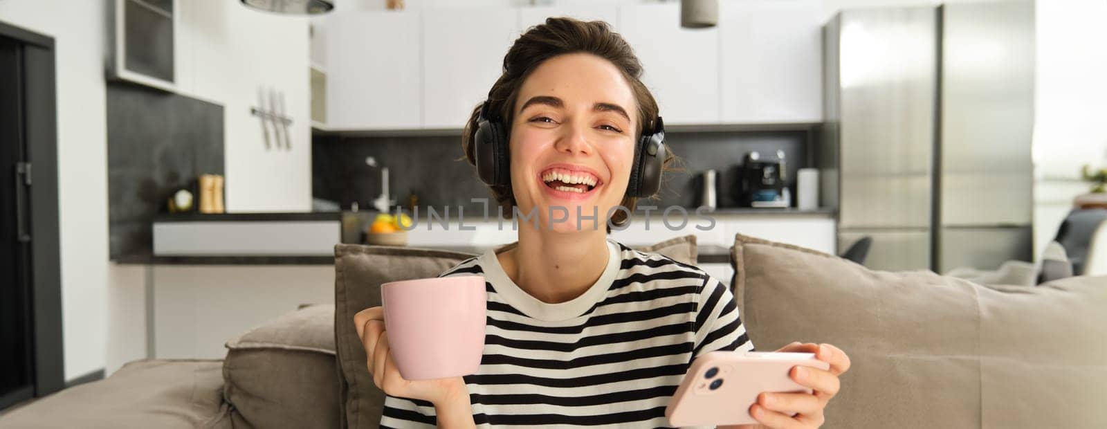 Close up portrait of happy woman with smartphone and headphones, drinking tea and watching videos on social media app, laughing and smiling by Benzoix