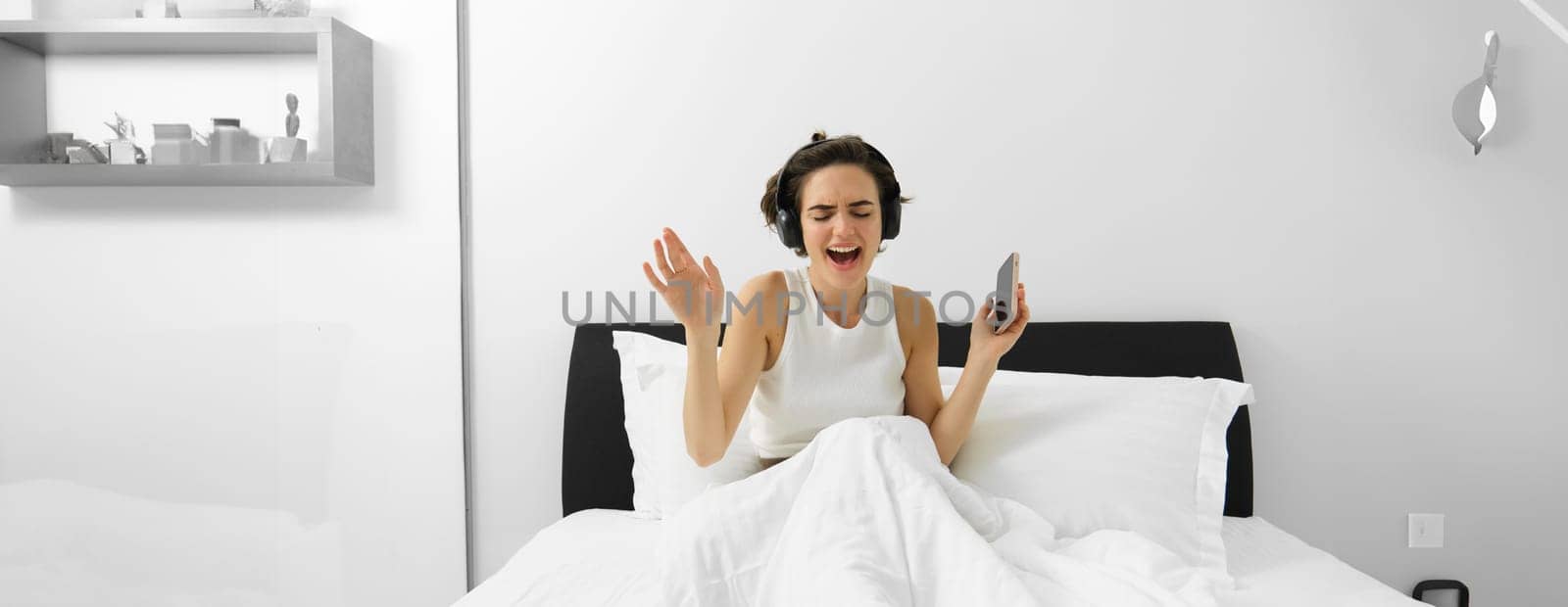 Portrait of happy woman singing in her bed, wakes up and listens to music in wireless headphones, holding mobile phone.