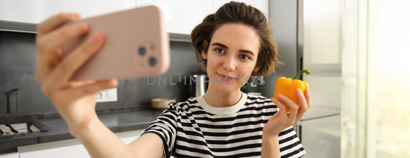 Close up portrait of cute brunette woman, taking selfie with yellow fresh pepper, recording video on smartphone, posing with vegetables in the kitchen, making cooking blog on social media app.