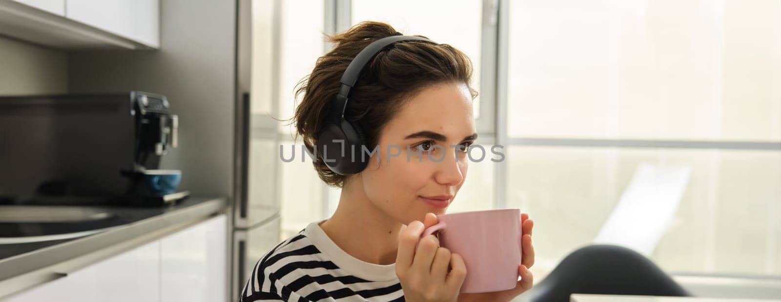 Close up portrait of smiling brunette woman, student drinks her tea and listens music or ebook in headphones, sits in kitchen and relaxes.