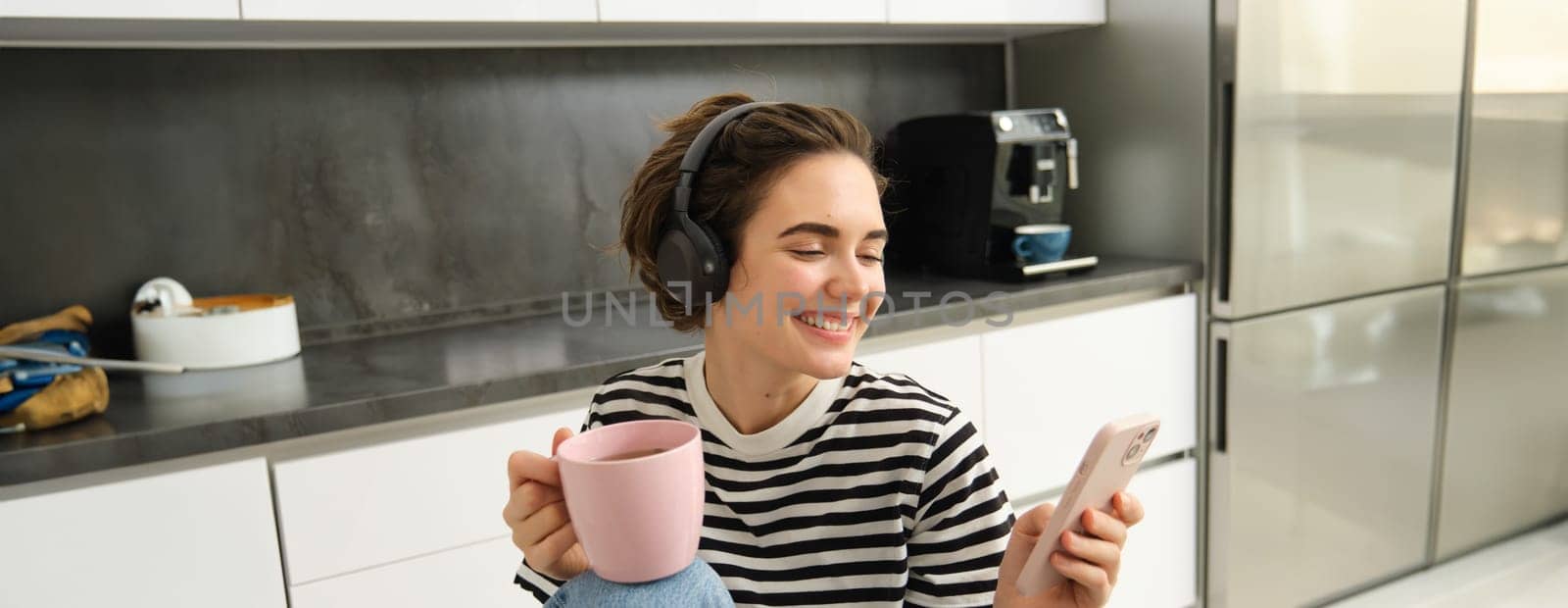 Portrait of smiling young woman, using mobile phone, sitting in kitchen and drinking hot tea, browsing social media and relaxing at home by Benzoix