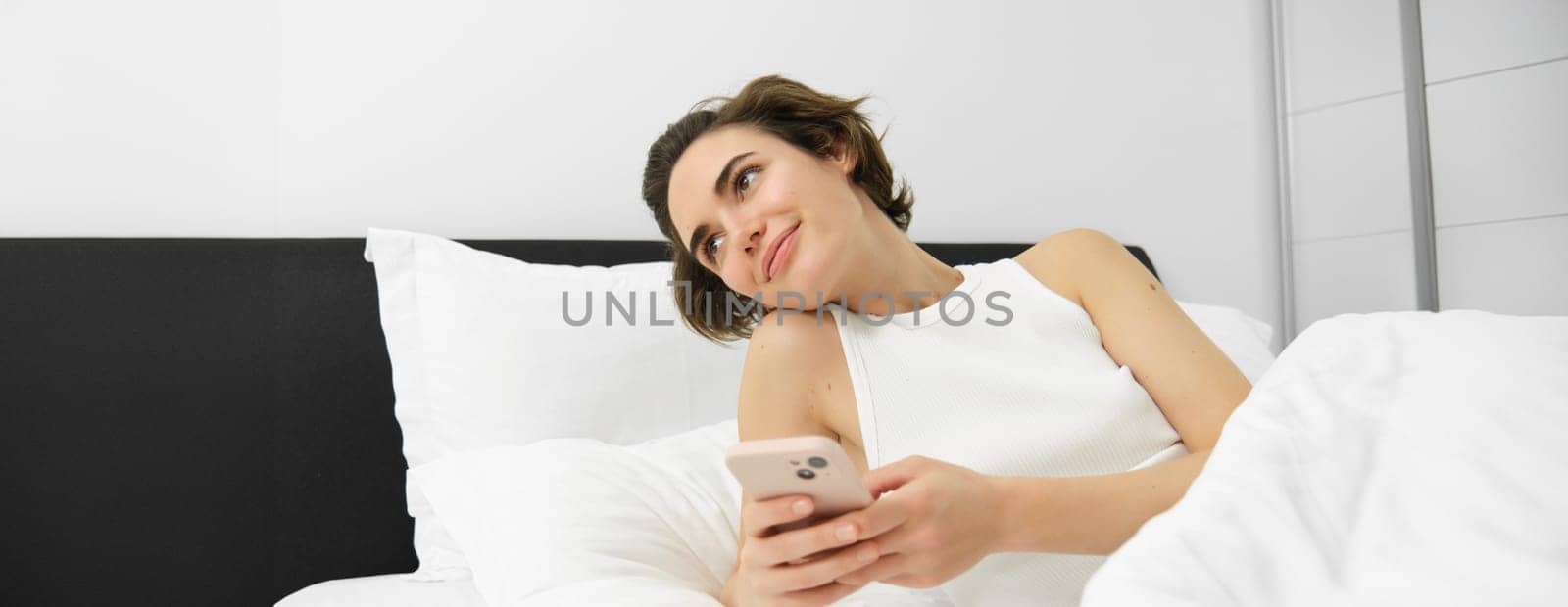 Portrait of woman lying in her bed, checking phone, sets up alarm clock to wake up in the morning.