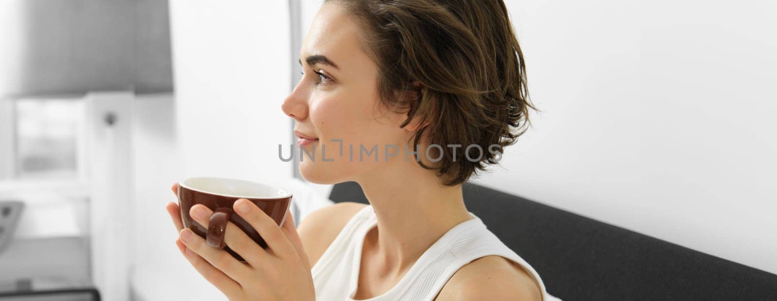 Sensual morning concept. Young beautiful woman drinking coffee in bed, holding mug and smelling nice aroma of freshly brewed drink by Benzoix