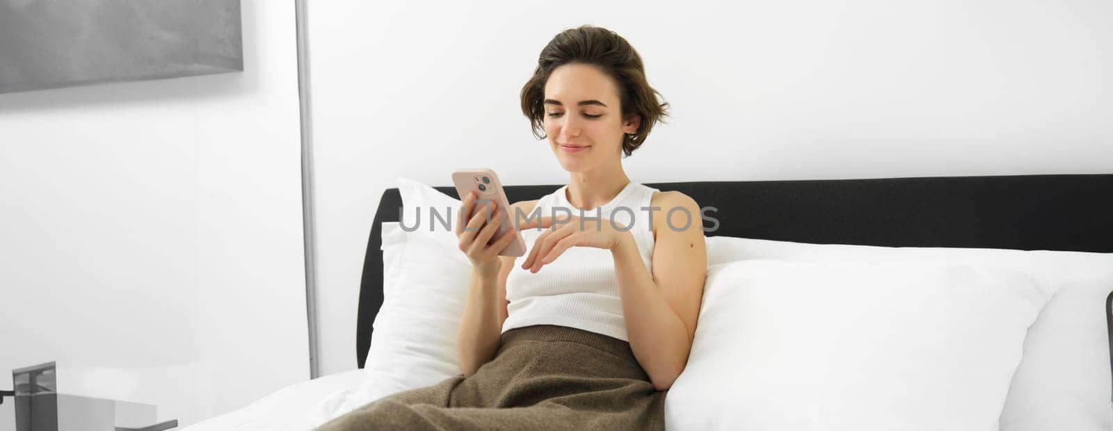 Portrait of cute female model in bedroom, holding phone, typing message on smartphone, resting in bed and smiling by Benzoix