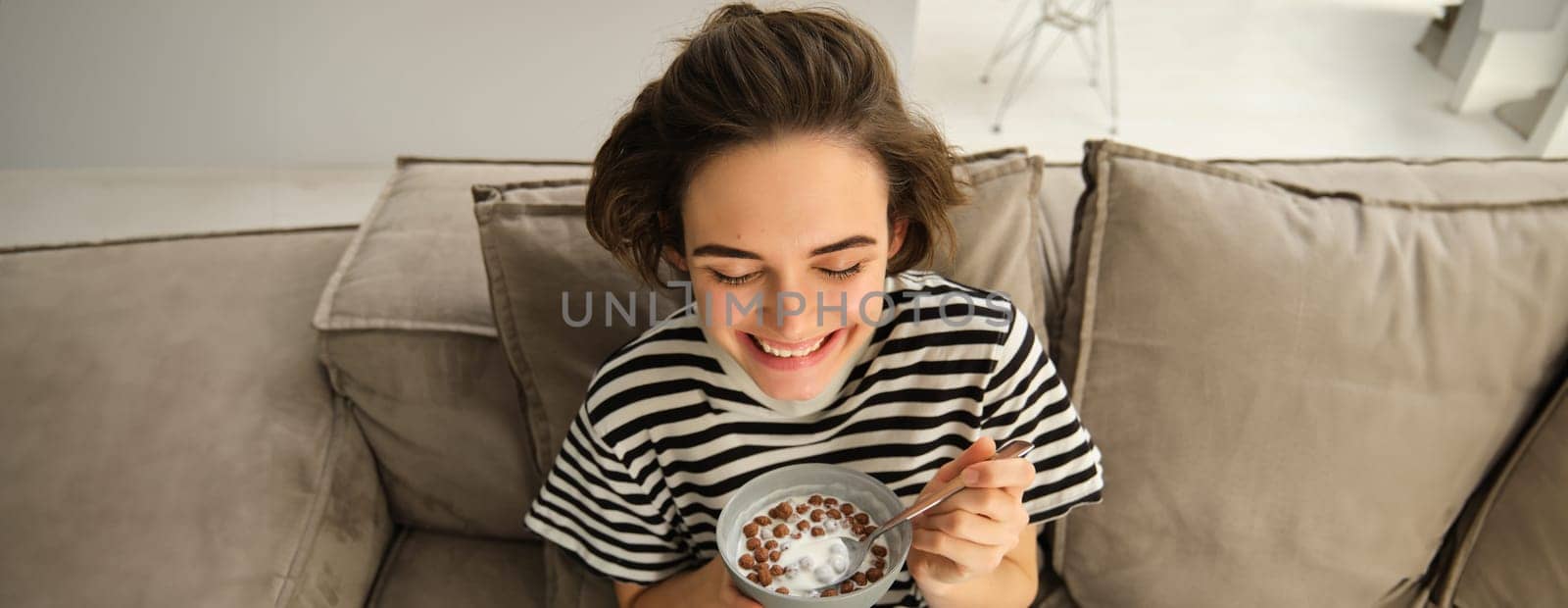 Close up of cute young female model, eating cereals with milk, enjoys her breakfast on sofa in living room, smiling and looking happy by Benzoix