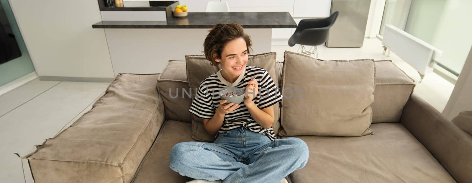 Portrait of happy brunette woman sitting on sofa and enjoying her breakfast, eating cereals, healthy granola in bowl, spending time in living room by Benzoix