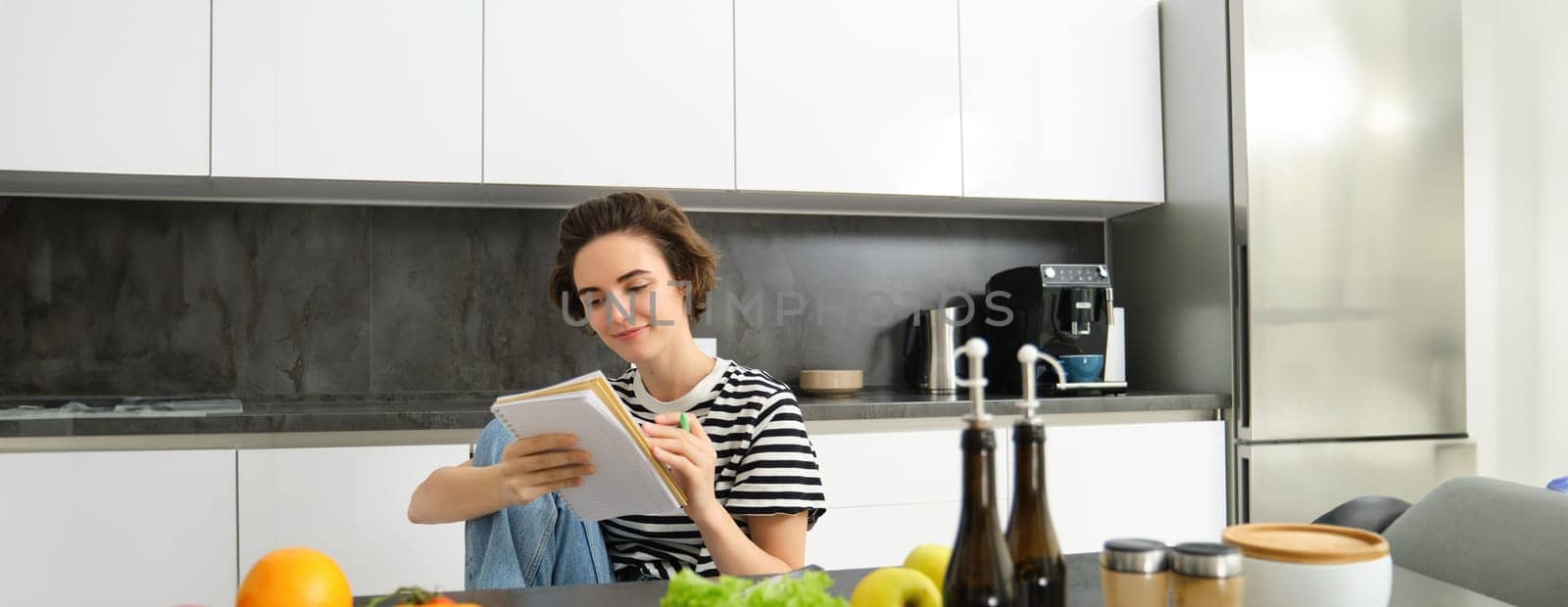 Portrait of young woman cooking, writing notes, grocery list in notebook, creating list of meals to cook through meal, sitting in kitchen near vegetables and chopping board by Benzoix