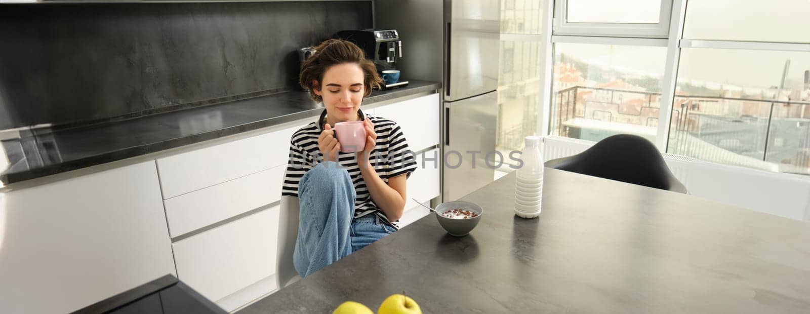 Cute brunette woman, student eating breakfast in morning, drinking hot tea, smiling and looking pleased by Benzoix