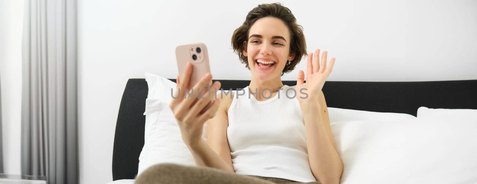 Cheerful smiling woman resting in bed, connects to online video call while lying in bedroom, talking to friend via smartphone app by Benzoix