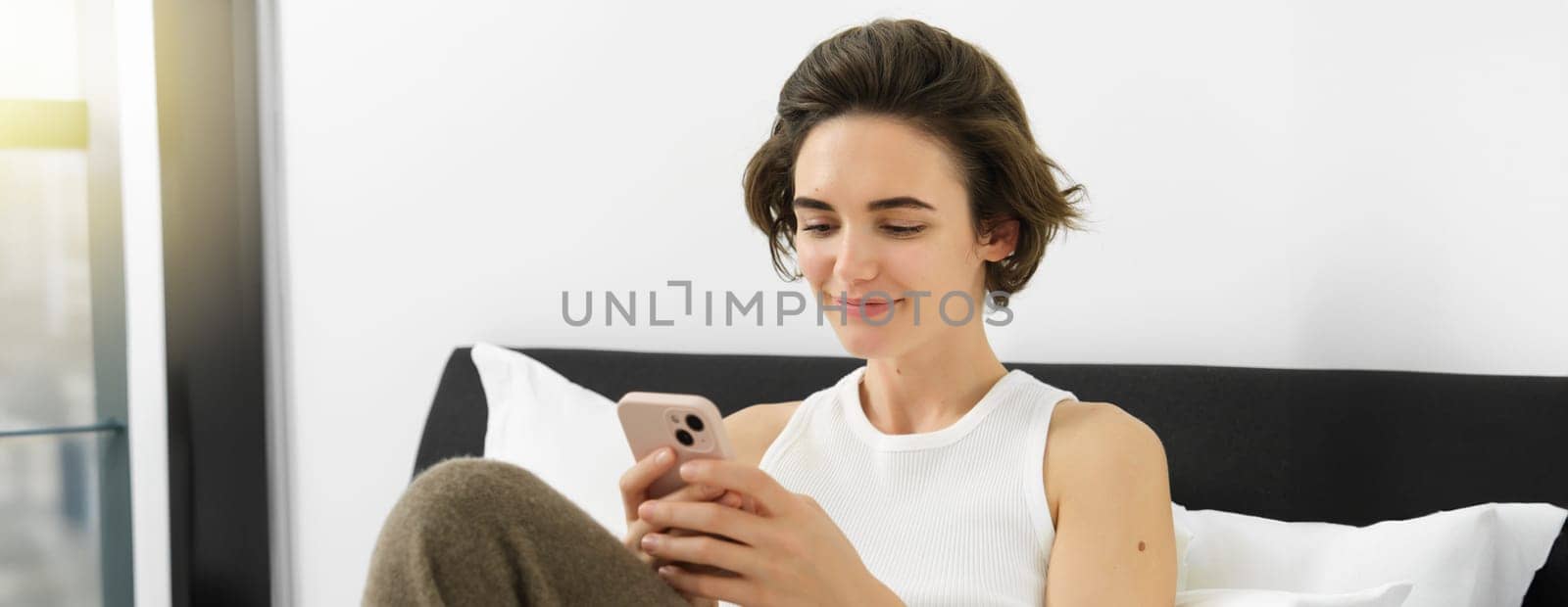 Portrait of young beautiful brunette woman, sitting on bed with smartphone, using mobile phone app, sending message and smiling, reading on telephone application in bedroom.