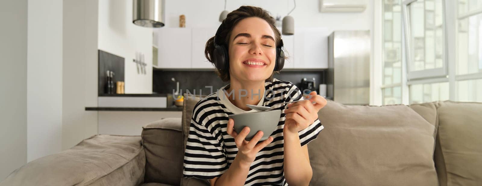 Portrait of woman laughing and smiling in headphones, eating cereals for breakfast and sitting on sofa in front of tv.