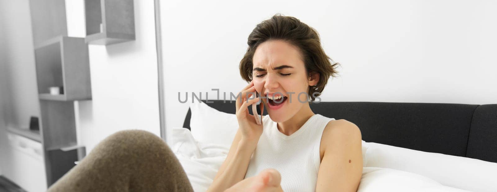 Young woman lying in bed, talking to friend over the phone, calling someone and complaining, gesturing with emotions, resting in bedroom.