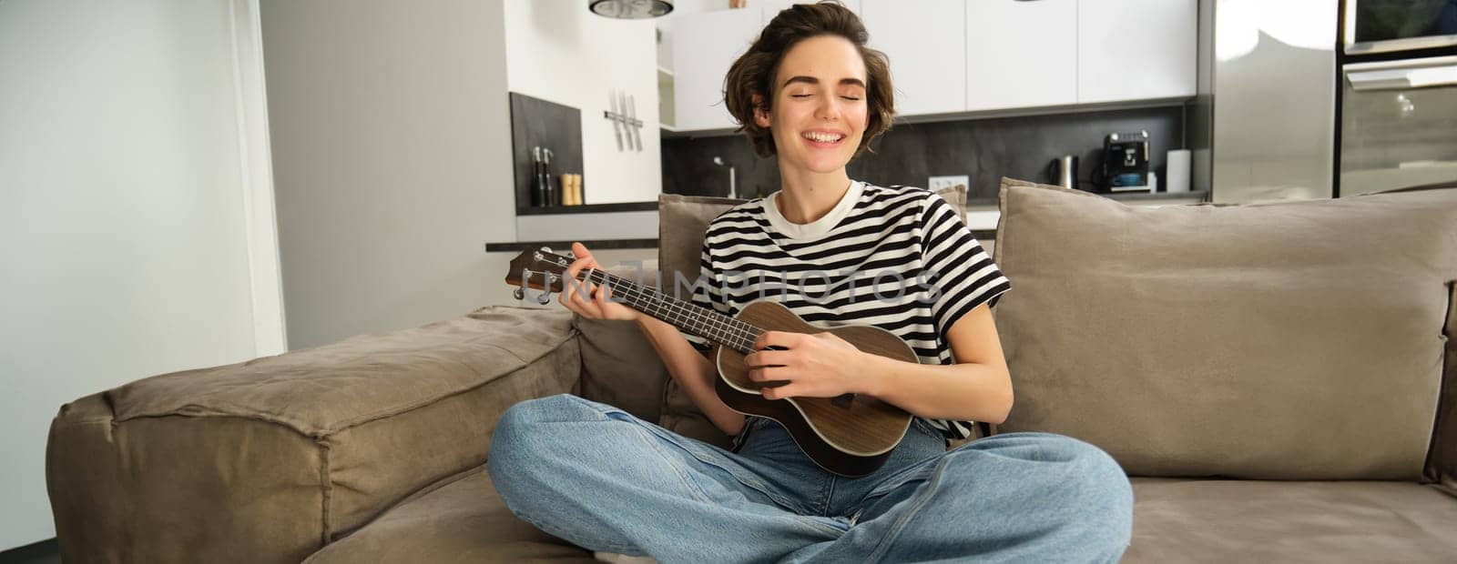 Portrait of cheerful young woman playing her ukulele, singing and laughing, sitting in living room at home by Benzoix